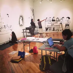 People work on murals and at tables in an art studio with drawings on the walls and supplies on the floor.