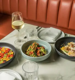 Three colorful dishes and a glass of white wine on a marble table with plates and napkins in a restaurant booth.