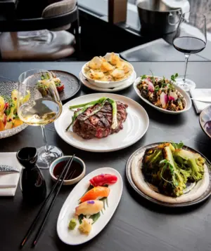 A table set with steak, sushi, vegetables, appetizers, and glasses of red and white wine.