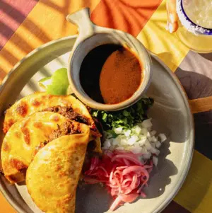Three birria tacos with cilantro, onions, pickled onions, lime, and consommé on a plate under sunlight.