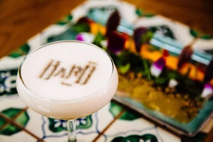 A cocktail with foam art in a glass, placed on a colorful tiled table next to a decorated plate.
