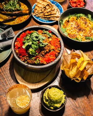 A table set with colorful rice, guacamole, chips, flatbread, curry, and a cocktail with a lemon slice.