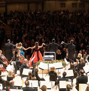 Five performers hold hands and bow to an applauding audience in a concert hall, with an orchestra in front.