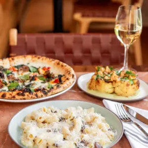 Plate of pasta, pizza, and roasted potatoes on a table with a glass of white wine and cutlery.