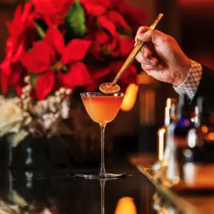 A hand stirs a cocktail in a glass, with red poinsettias and festive decor in the background.