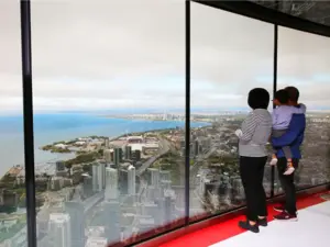 Two people and a child look out a large window at a city skyline and waterfront from a high vantage point.