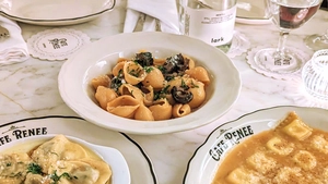 A bowl of shell pasta with mushrooms sits on a marble table beside plates of ravioli and drinks.