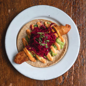 A fish taco topped with red cabbage slaw, sauce, and cilantro on a white plate, on a wooden table.