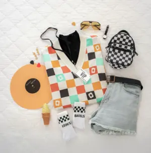 Colorful checkered sweater, denim shorts, checkered socks, mini backpack, sunglasses, and accessories on a bed.