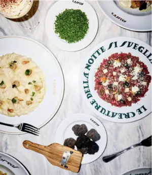 Plates of risotto, steak tartare, herbs, and truffles on a white marble table seen from above.