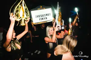 Women in a nightclub hold up bottles, lights, and a sign that reads 'OVESUSNOT IYKYK.