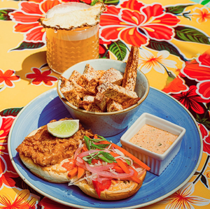 A fried chicken sandwich with pickled veggies, fries, dip, and a drink on a colorful floral tablecloth.