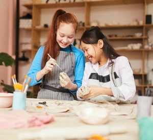 Two girls smiling and making pottery together in a bright, creative studio with art supplies.