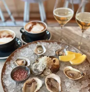 A plate of oysters on ice with lemon wedges, two glasses of wine, and two cups of coffee on a table.