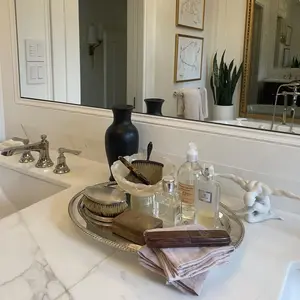 A bathroom counter with a silver tray holding soap, bottles, towels, and a black vase beside a sink.