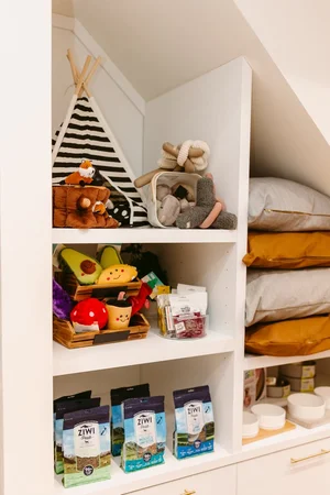 White shelves holding pet supplies, toys, treats, and bedding under a slanted ceiling in a tidy space.