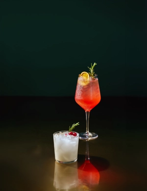 Two cocktails on a dark surface: one in a wine glass with garnish, one in a short glass with ice and a cherry.