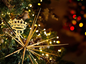 Gold crown and star ornaments hang on a Christmas tree, with white lights and blurred lights in the background.