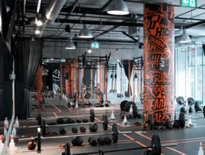 Spacious gym with weights, kettlebells, orange pillars with graffiti, and workout stations.
