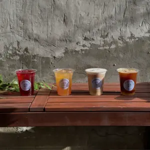 Four different iced drinks in plastic cups on a wooden table against a textured gray wall with some sunlight.