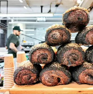 Stacked poppy seed-covered pastries on a wooden counter with a barista working in the background.