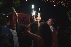 People at a dimly lit party raise bottles and glowing lights in celebration under twinkling string lights.