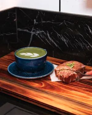 A blue cup of matcha latte with latte art next to a sliced pastry on a wooden tray.