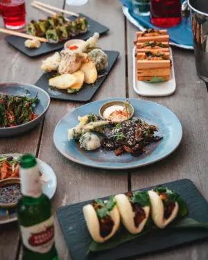 A table with assorted Asian dishes, bao buns, tempura, and drinks arranged on plates and boards.