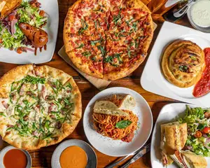 An overhead view of pizzas, salad, pasta, and sauces arranged on a wooden table.