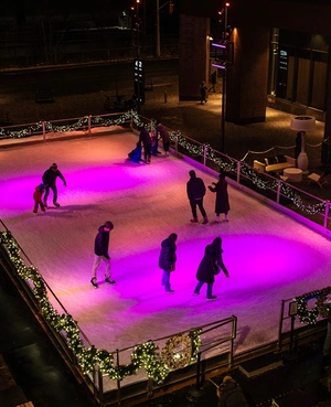 People ice skating at night on a rink lit with purple lights and decorated with holiday garlands.