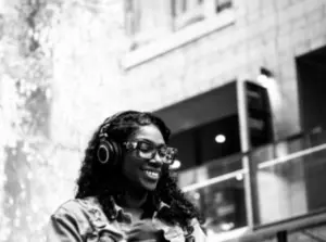Smiling woman wearing headphones and glasses stands outdoors in an urban setting, holding a smartphone.