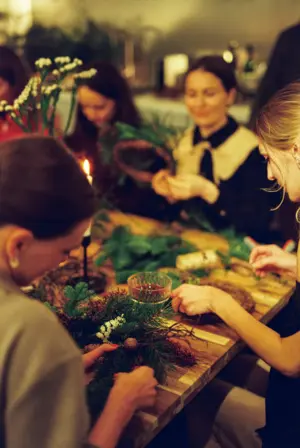 Four women sit around a table making festive wreaths with greenery and pinecones in a cozy, softly lit room.