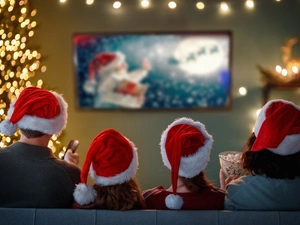 Four people in Santa hats watch a Christmas movie on TV, with popcorn and holiday lights in the background.