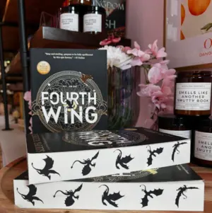 A stack of 'Fourth Wing' books with dragon designs, displayed with flowers and scented candles.