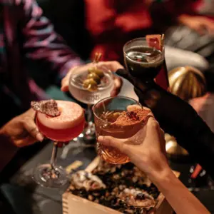 Four people clink cocktail glasses together over a table with food, celebrating in a festive atmosphere.