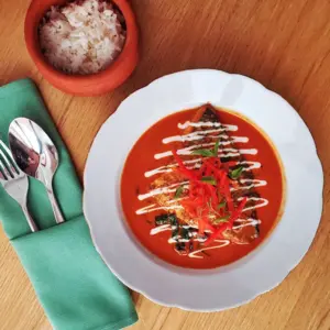 White plate of red curry with garnish, next to a bowl of rice and utensils on a green napkin.