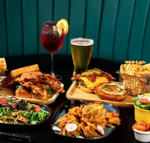 A variety of burgers, fries, salads, fried appetizers, a beer, and a red cocktail on a restaurant table.
