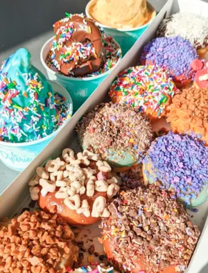 Box of colorful donuts with various toppings beside cups of brightly decorated ice cream with sprinkles.