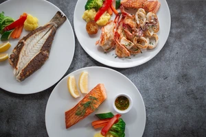 Three plates with grilled fish, seafood, vegetables, lemon wedges, and dipping sauce on a gray surface.