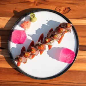 Artfully plated sushi rolls with sauces, wasabi, and pickled ginger on a white plate atop a wooden table.