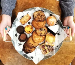 A person holds a tray filled with assorted cookies, muffins, and pastries on star-patterned paper.