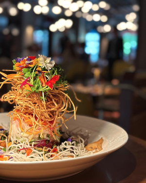 A vibrant salad topped with crispy noodles and edible flowers, served on a white plate in a softly lit restaurant.