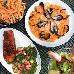 Plates of seafood pasta, fries, grilled salmon with salad, and shrimp cocktail on a metal table.