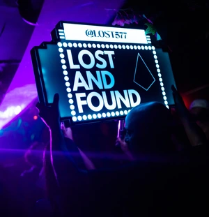 Person holding a brightly lit 'Lost and Found' sign in a dark, neon-lit club setting.