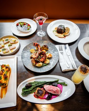 A table set with various gourmet dishes, cocktails, and neatly arranged silverware on a wooden surface.