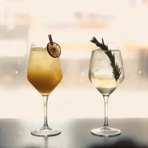 Two cocktails in stemmed glasses; one with an orange slice, the other with rosemary garnish, on a table.