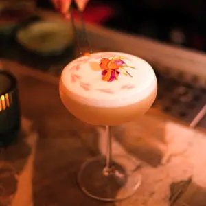 A cocktail with a foamy top, decorated with pink flower petals, in a coupe glass on a marble surface.