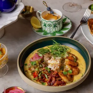 A colorful bowl of creamy soup with grilled chicken, vegetables, and herbs, served with drinks on a table.