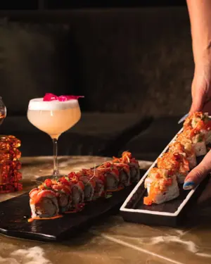 Two sushi rolls on black plates with a hand serving one, and a cocktail garnished with a flower in the background.
