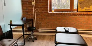 A tattoo studio with a brick wall, window, desk, stool, and black tattoo beds.